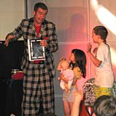Magician on the left pulling a face at two children who are standing next to him on the stage and looking up at him.